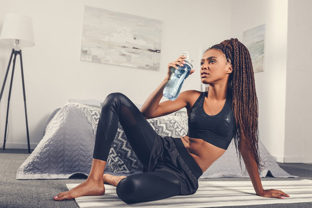 sportive african american woman drinking water after workout at home on yoga mat